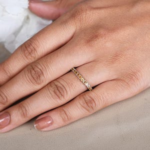 May include: A gold ring set with small, evenly spaced yellow gemstones. The ring is on a person's finger, with the hand resting on a neutral-toned surface. White flowers are in the background.