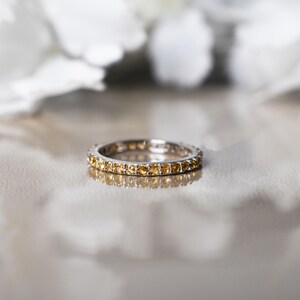 May include: A silver ring set with small, round, yellow gemstones. The ring sits on a reflective surface, with blurred white shapes in the background. The ring's design is simple and elegant, suitable for everyday wear.