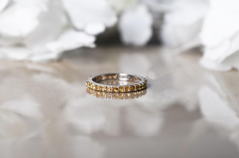 May include: A silver ring set with small, round, yellow gemstones. The ring sits on a reflective surface, with blurred white shapes in the background. The ring's design is simple and elegant, suitable for everyday wear.