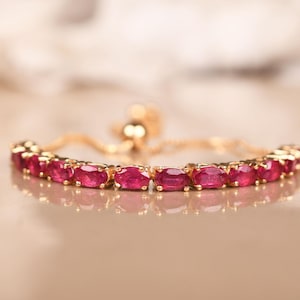 Puede incluir: Una pulsera dorada con una serie de piedras preciosas rojas ovaladas y vibrantes. La pulsera tiene una cadena delicada y un cierre deslizante para un ajuste ajustable. Las piedras están engastadas en biseles de oro individuales.