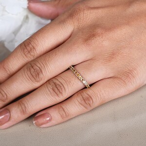 May include: A gold ring set with small yellow gemstones. The ring is worn on a finger, with the hand resting on a neutral surface. The nails are painted a neutral colour. White flowers are in the background.