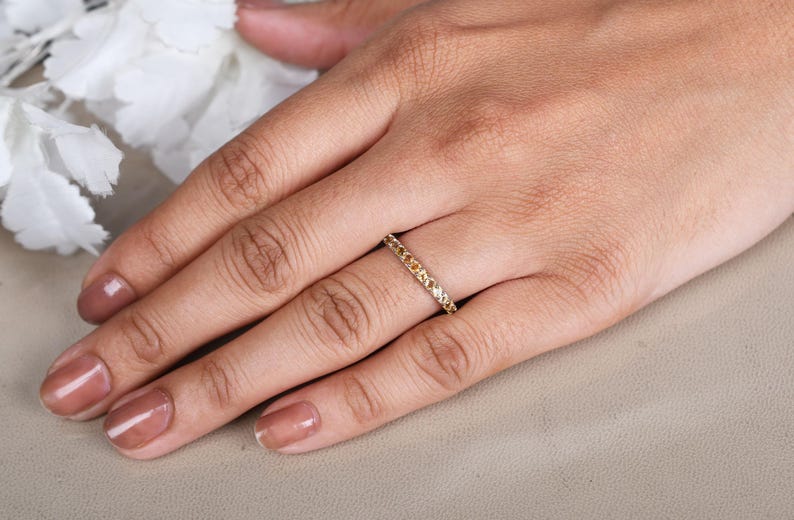 May include: A gold ring set with small yellow gemstones. The ring is worn on a finger, with the hand resting on a neutral surface. The nails are painted a neutral colour. White flowers are in the background.