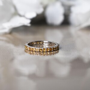 May include: A silver ring featuring a continuous line of small, yellow gemstones. The ring is set against a reflective surface, with blurred white flowers in the background. The gemstones are evenly spaced around the band.
