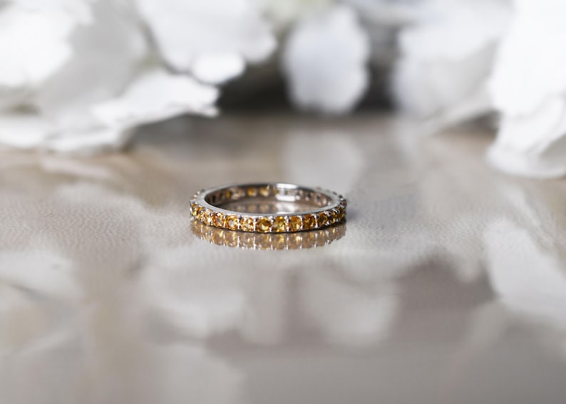 May include: A silver ring featuring a continuous line of small, yellow gemstones. The ring is set against a reflective surface, with blurred white flowers in the background. The gemstones are evenly spaced around the band.