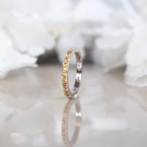 May include: A silver ring featuring a continuous row of small, round, yellow gemstones. The ring is reflected on a surface, with a blurred white backdrop. The ring is likely a wedding band or eternity ring.