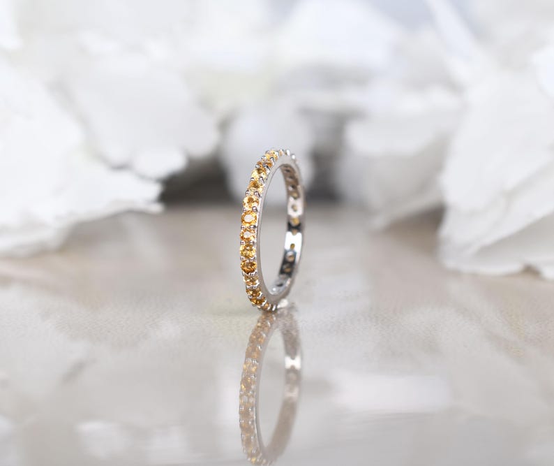 May include: A silver ring featuring a continuous row of small, round, yellow gemstones. The ring is reflected on a surface, with a blurred white backdrop. The ring is likely a wedding band or eternity ring.