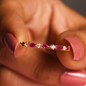 Peut inclure: Une bague en or ornée de pierres précieuses alternant rubis et diamants. La bague présente des rubis taille marquise et des diamants ronds sertis dans une fine monture. La bague est tenue entre les doigts avec du vernis à ongles rose.