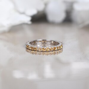 May include: A silver ring featuring a continuous band of small, round, yellow gemstones. The ring is set against a soft, blurred background. The ring is a piece of jewellery.