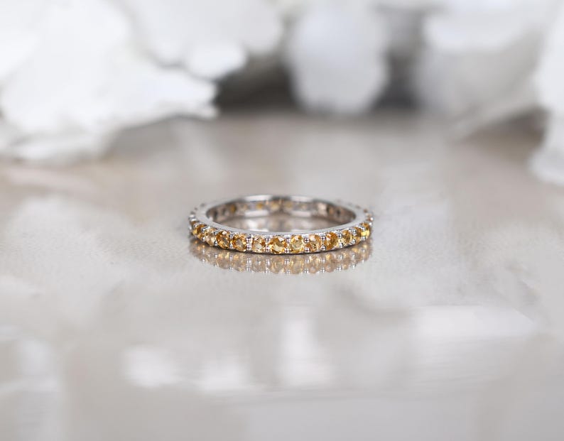 May include: A silver ring featuring a continuous band of small, round, yellow gemstones. The ring is set against a soft, blurred background. The ring is a piece of jewellery.