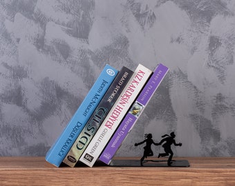 Puede incluir: Un sujetalibros de metal negro con dos siluetas de personas corriendo. El sujetalibros sostiene una pila de cinco libros con títulos visibles: "DÜŞLER KRALLIĞI", "OK GISELLE GREEN CILSICD", "BRACO STOKER HAYATA UYANMAK", "KIZ KARDEŞİN HEDİYESİ", y "MARY E. PEARSON".