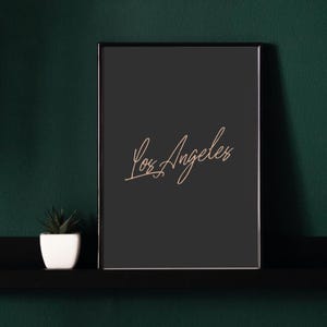 May include: A black framed print with the text "Los Angeles" written in a cursive script in a light brown color.