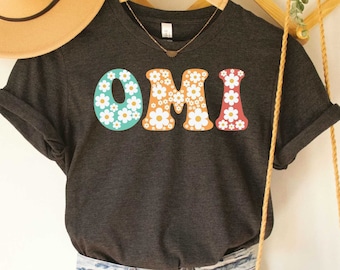 May include: Dark grey heather V-neck t-shirt with the word "OMI" in large, colorful letters decorated with white daisies. The letters are outlined in red, teal, and orange.