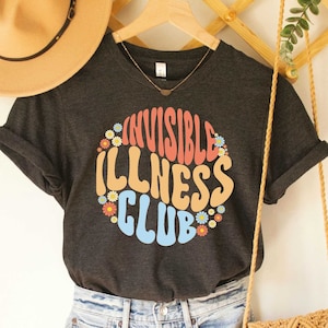 K&ouml;nnte beinhalten: Dunkelgraues T-Shirt mit einem bunten Aufdruck, auf dem "Invisible Illness Club" steht, umgeben von Blumen.