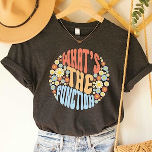 May include: Dark grey heather t-shirt with the text "WHAT'S THE FUNCTION" in a colorful, retro-style design. The design is a circle filled with daisies. The shirt is hanging on a wooden hanger.