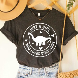 Puede incluir: Camiseta de color gris oscuro jaspeado con un gr&aacute;fico blanco de un dinosaurio y el texto "Just a girl who loves dinosaurs".