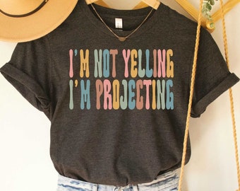 May include: Dark grey heather V-neck t-shirt with the text "I'm not yelling I'm projecting" in colourful retro font.
