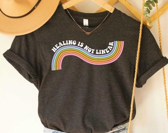May include: Dark grey heather t-shirt with a rainbow graphic and the text "Healing is not linear".