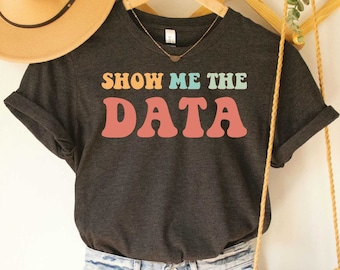 May include: Dark grey heather t-shirt with the text "SHOW ME THE DATA" in rainbow colors. The shirt is being modeled with a straw hat and a crocheted bag.