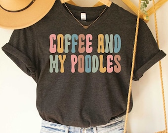 Peut inclure: T-shirt en jersey gris foncé chiné avec le texte "Coffee and My Poodles" en lettres rétro colorées. Le t-shirt est porté avec un chapeau de paille et un sac en crochet.