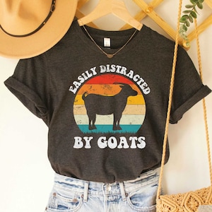 May include: Dark grey heather V-neck t-shirt with a vintage-style graphic of a goat in front of a sunset. The text "Easily Distracted By Goats" is printed on the shirt.