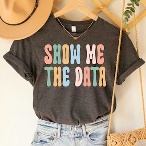May include: A dark grey heather t-shirt with the text "Show Me The Data" in colorful retro font.