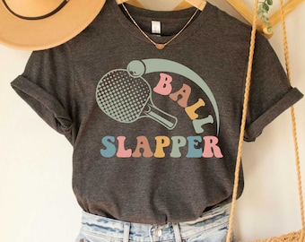 May include: Dark grey heather t-shirt with a retro graphic of a ping pong paddle and ball with the text "Ball Slapper".