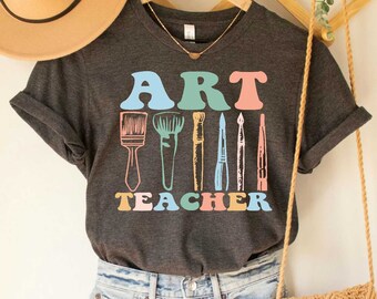 Peut inclure: T-shirt à col en V gris foncé chiné avec le texte "ART TEACHER" et un graphique coloré de pinceaux et de crayons.