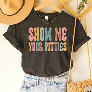 May include: Dark grey heather t-shirt with the text "Show Me Your Pitties" in rainbow colors. The shirt is shown on a model wearing denim shorts and a straw hat.