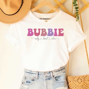 May include: White t-shirt with the text "BUBBIE" in a rainbow gradient font. The text "Wesley Bennet Alina" is written in cursive below the word "BUBBIE".