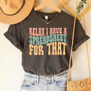 May include: A dark gray t-shirt with the text "Relax I have a spreadsheet for that" in a retro font with pink, blue, and orange colors.
