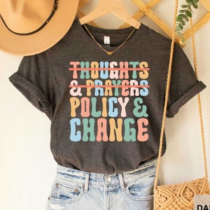 May include: Dark grey heather t-shirt with the text "Thoughts & Prayers" crossed out and replaced with "Policy & Change" in colorful retro font.