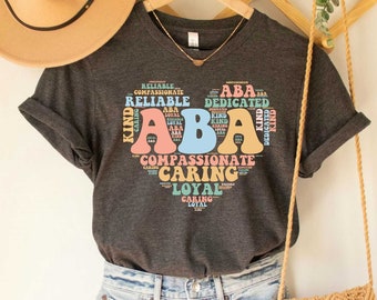 May include: A dark gray t-shirt with a graphic design of the letters "ABA" in a heart shape. The letters are surrounded by words describing positive personality traits, such as "reliable", "kind", "compassionate", and "caring".