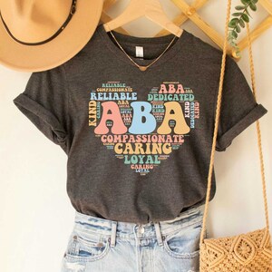 May include: A dark gray t-shirt with a graphic design of the letters "ABA" in a heart shape. The letters are surrounded by words describing positive personality traits, such as "reliable", "kind", "compassionate", and "caring".