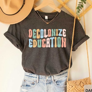 May include: A dark grey heather t-shirt with the text "DECOLONIZE EDUCATION" in rainbow colored letters. The shirt is being modeled on a person wearing denim shorts and a straw hat.