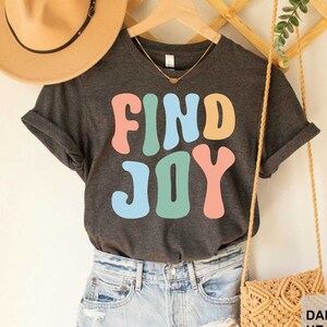 May include: A dark grey heather t-shirt with the text "FIND JOY" in colorful retro lettering.