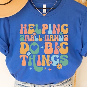 May include: Royal blue t-shirt with the text "HELPING SMALL HANDS DO BIG THINGS" in retro-style lettering. The design includes flowers, hearts, and stars in orange, green, and blue. The shirt is paired with denim shorts.
