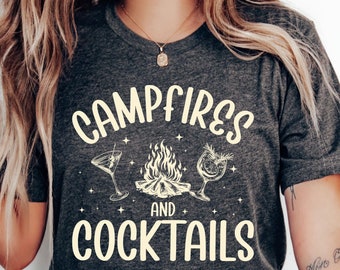 May include: A dark grey heather t-shirt with the text "Campfires and Cocktails" in a vintage-inspired font. The design features a campfire with two cocktails on either side.