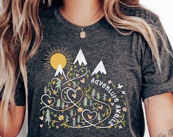 May include: A dark grey heather t-shirt featuring a graphic design of mountains, a sun, hearts, and trees. The text "Adventure Awaits" is written on the shirt.