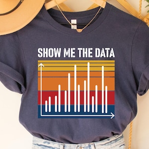 May include: Navy blue t-shirt with the text "SHOW ME THE DATA" above a graphic of a bar chart in red, orange, yellow, and blue. The shirt is a casual style.