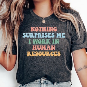 May include: A dark grey heather t-shirt with the text "NOTHING SURPRISES ME I WORK IN HUMAN RESOURCES" in colorful retro font.