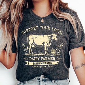 May include: A dark grey heather t-shirt with a white graphic of a cow and the text "Support Your Local Dairy Farmer - Fresh Milk Daily - Always on Tap".