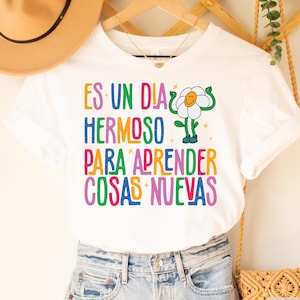 May include: White t-shirt with a colorful graphic of a flower with the text "ES UN DIA HERMOSO PARA APRENDER COSAS NUEVAS".