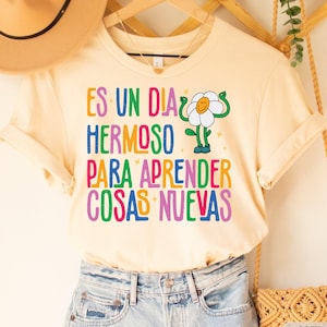 May include: A cream colored t-shirt with a colorful graphic that says "Es un dia hermoso para aprender cosas nuevas" and a cartoon flower with arms and legs.