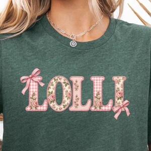 May include: A heather forest green short-sleeve t-shirt featuring the word LOLLI in large block letters. The letters have a pink and white gingham pattern with floral accents and are decorated with small pink bows on the ends.