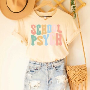 May include: A light beige t-shirt with the text "SCHOOL PSYCH" in colorful retro font.