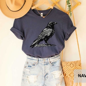 May include: A navy blue t-shirt with a black and white illustration of a raven. The t-shirt is shown on a model wearing denim shorts and a straw hat.