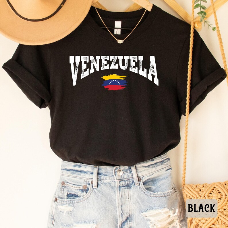 May include: Black t-shirt with white text that says "VENEZUELA" and a small Venezuelan flag graphic. The shirt is being modeled on a person wearing denim shorts.