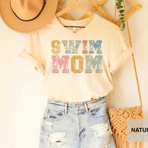 Pu&ograve; includere: Una maglietta beige chiaro con il testo "SWIM MOM" in lettere colorate e consumate. La maglietta &egrave; indossata con pantaloncini di jeans e una borsa di paglia.