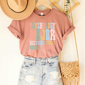 May include: A pink t-shirt with the text "Psychology Major Cuz Every Family Needs A Therapist" in colorful, retro-style lettering.
