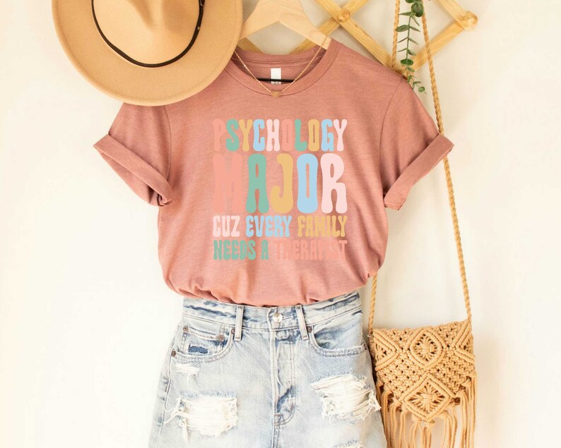 May include: A pink t-shirt with the text "Psychology Major Cuz Every Family Needs A Therapist" in colorful, retro-style lettering.